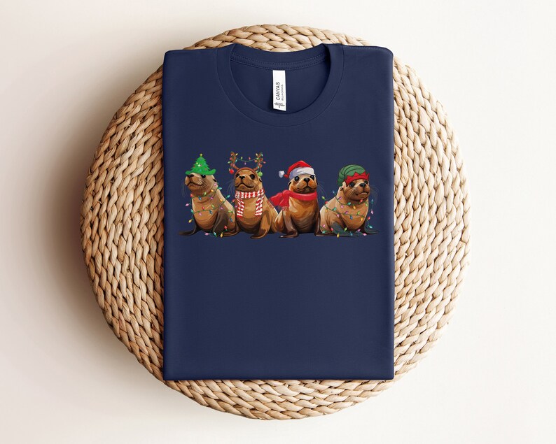 May include: A navy blue t-shirt with a graphic of four seals wearing Christmas hats and decorations. The seals are wearing a Santa hat, a green elf hat, a red and white Santa hat, and a green elf hat with a red pom-pom.