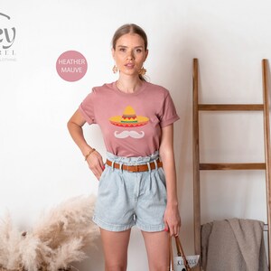 May include: A heather mauve t-shirt with a graphic of a sombrero and a mustache. The text "HEATHER MAUVE" is visible in the top left corner of the image.