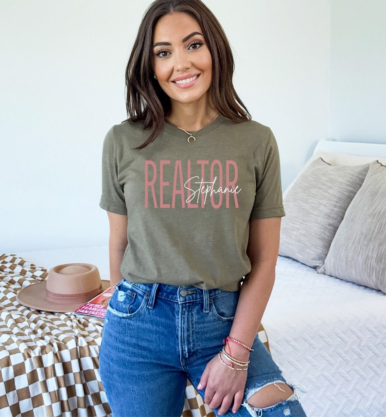 Peut inclure: Une femme portant un t-shirt vert avec le texte "REALTOR Stephanie" en lettres roses. Elle porte un jean bleu et a de longs cheveux bruns.