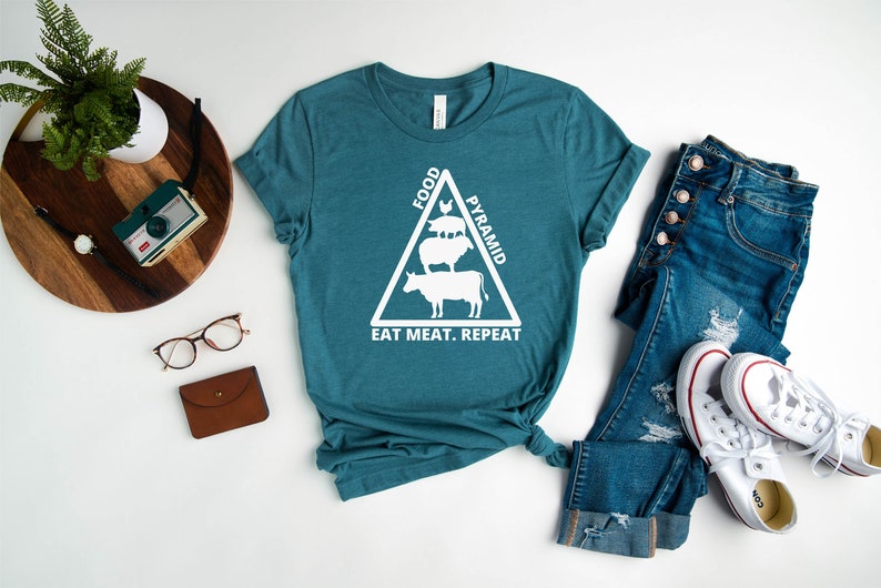 May include: Teal blue t-shirt with a white graphic of a food pyramid made of animals. The text "FOOD PYRAMID" is above the animals and the text "EAT MEAT. REPEAT" is below the animals.