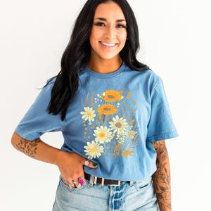 K&ouml;nnte beinhalten: Ein blaues T-Shirt mit einem Vintage-Blumendruck mit gelben und wei&szlig;en G&auml;nsebl&uuml;mchen und braunen Stielen. Das Shirt hat einen Rundhalsausschnitt und kurze &Auml;rmel.