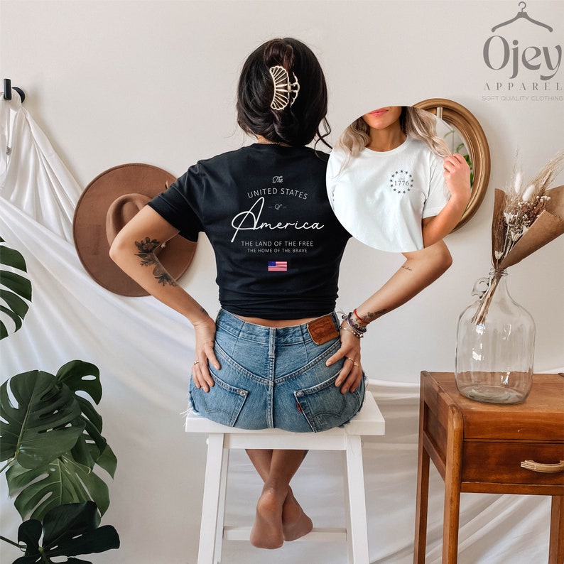 Peut inclure: Un t-shirt noir avec un motif blanc qui dit "The United States of America - The Land of the Free - The Home of the Brave" avec un petit drapeau am&eacute;ricain. Le t-shirt est port&eacute; par une personne avec une pince &agrave; cheveux dor&eacute;e.