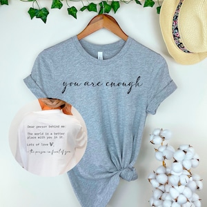May include: A gray t-shirt with the text "you are enough" printed on the front. The back of the shirt has the text "Dear person behind me: The world is a better place with you in it. Lots of love, -the person in front of you".