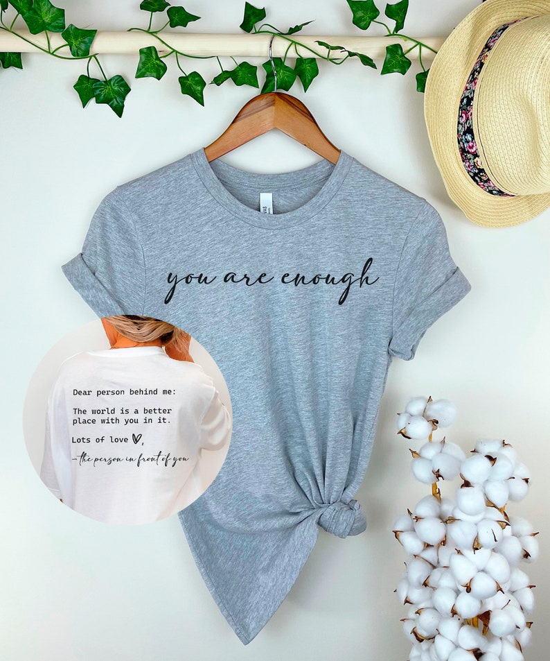 May include: A gray t-shirt with the text "you are enough" printed on the front. The back of the shirt has the text "Dear person behind me: The world is a better place with you in it. Lots of love, -the person in front of you".