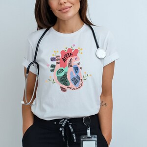 May include: White t-shirt with a colorful anatomical heart illustration. The heart is labeled with different parts, including the aorta, myocardium, and ventricles. The shirt has a stethoscope around the neck.