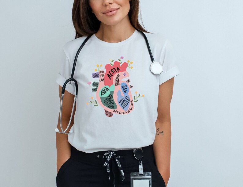 May include: White t-shirt with a colorful anatomical heart illustration. The heart is labeled with different parts, including the aorta, myocardium, and ventricles. The shirt has a stethoscope around the neck.