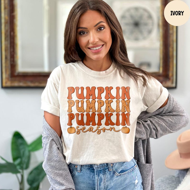 May include: Ivory-colored t-shirt with the words "Pumpkin Season" in orange and brown lettering. The word "Pumpkin" is repeated three times, with the middle one having a leopard print design. Two small pumpkins are on the bottom.