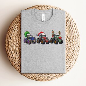 Peut inclure: T-shirt gris avec un motif de trois tracteurs d&eacute;cor&eacute;s pour No&euml;l. Les tracteurs sont bleu, rouge et vert, et chacun a une d&eacute;coration de No&euml;l diff&eacute;rente. Le tracteur bleu a un chapeau de P&egrave;re No&euml;l vert, le tracteur rouge a un chapeau de P&egrave;re No&euml;l rouge et le tracteur vert a des lumi&egrave;res de No&euml;l.