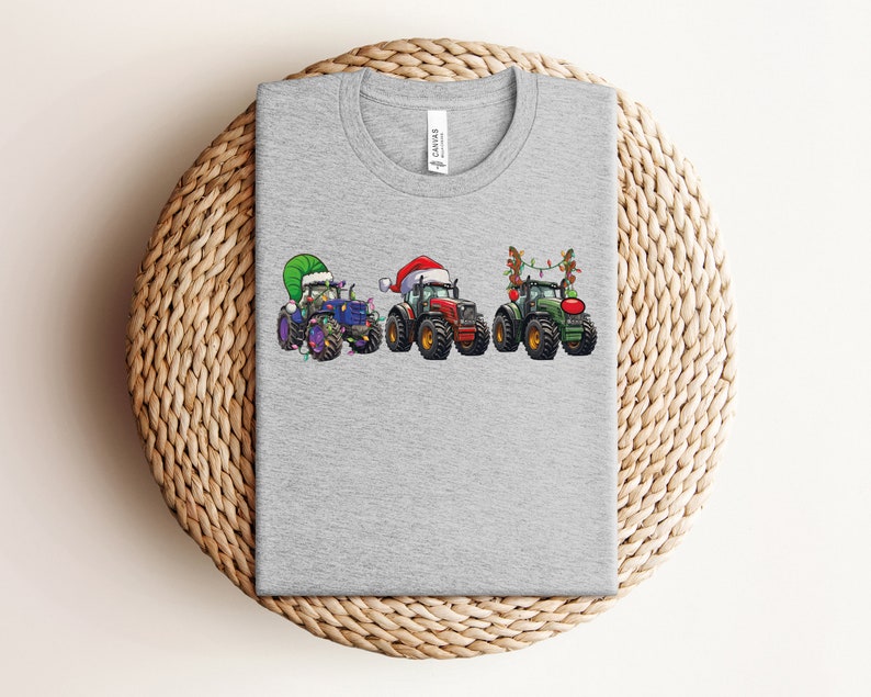 Peut inclure: T-shirt gris avec un motif de trois tracteurs d&eacute;cor&eacute;s pour No&euml;l. Les tracteurs sont bleu, rouge et vert, et chacun a une d&eacute;coration de No&euml;l diff&eacute;rente. Le tracteur bleu a un chapeau de P&egrave;re No&euml;l vert, le tracteur rouge a un chapeau de P&egrave;re No&euml;l rouge et le tracteur vert a des lumi&egrave;res de No&euml;l.