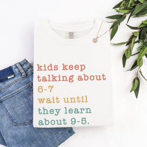 May include: A white t-shirt with the text "kids keep talking about 6-7 wait until they learn about 9-5." The text is in various colours. Blue jeans are folded next to the shirt. A gold necklace and greenery are also in the image.