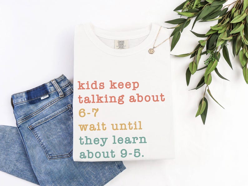 May include: A white t-shirt with the text "kids keep talking about 6-7 wait until they learn about 9-5." The text is in various colours. Blue jeans are folded next to the shirt. A gold necklace and greenery are also in the image.