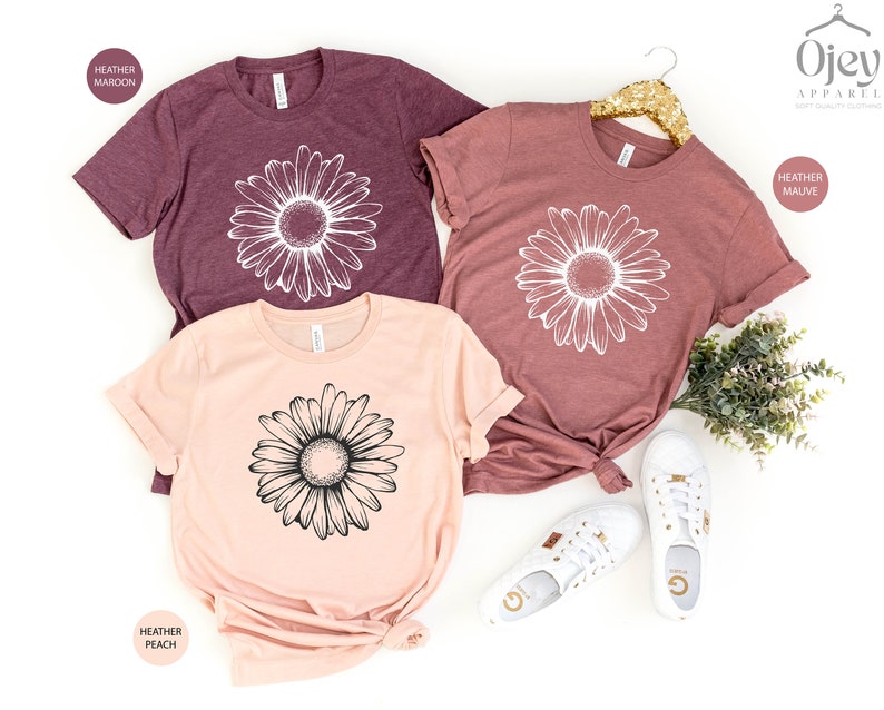 Puede incluir: Tres camisetas con un dise&ntilde;o gr&aacute;fico de margarita. Las camisetas son de color marr&oacute;n jaspeado, malva jaspeado y melocot&oacute;n jaspeado.