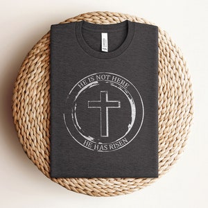 K&ouml;nnte beinhalten: Ein dunkelgraues T-Shirt mit einem wei&szlig;en Kreisdesign, das ein Kreuz und den Text "Er ist nicht hier, Er ist auferstanden" zeigt.