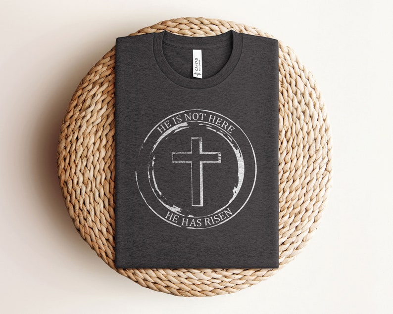 K&ouml;nnte beinhalten: Ein dunkelgraues T-Shirt mit einem wei&szlig;en Kreisdesign, das ein Kreuz und den Text "Er ist nicht hier, Er ist auferstanden" zeigt.