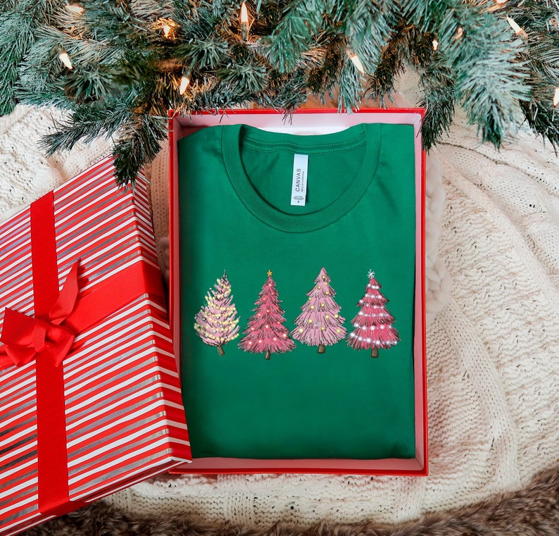 May include: A green t-shirt featuring a pink and white Christmas tree design. The shirt is folded and placed in a red and white striped gift box.