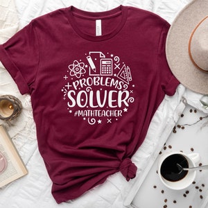 May include: A maroon t-shirt with a white graphic design that says "PROBLEMS? SOLVER #MATHTEACHER". The graphic includes a calculator, pencil, ruler, protractor, and atom symbol.