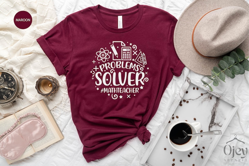 May include: A maroon t-shirt with a white graphic design that says "PROBLEMS? SOLVER #MATHTEACHER". The graphic includes a calculator, pencil, ruler, protractor, and atom symbol.