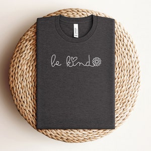 Puede incluir: Una camiseta gris oscuro con el texto "be kind" en blanco y un peque&ntilde;o gr&aacute;fico de margarita.