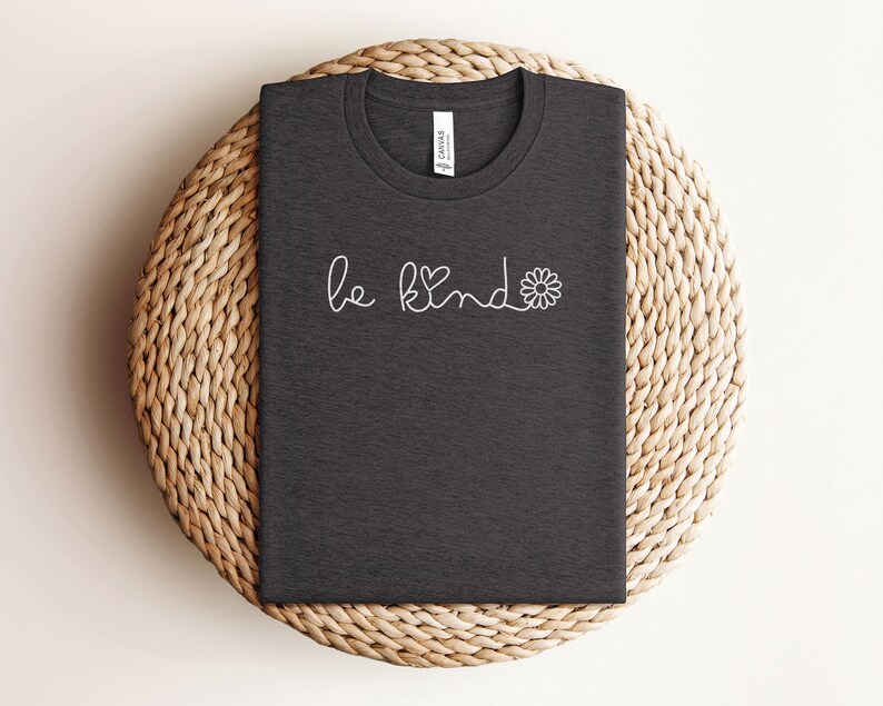 Puede incluir: Una camiseta gris oscuro con el texto "be kind" en blanco y un peque&ntilde;o gr&aacute;fico de margarita.