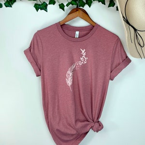 Op de afbeelding: Een mauvekleurig T-shirt met een witte afbeelding van een veer met vogels die eromheen vliegen.