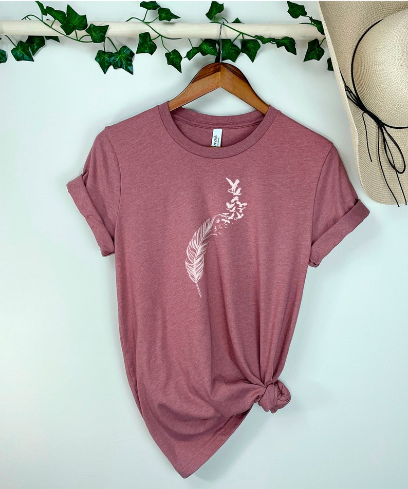 Op de afbeelding: Een mauvekleurig T-shirt met een witte afbeelding van een veer met vogels die eromheen vliegen.