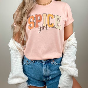 May include: A pink t-shirt with the text "SPICE girl" in a retro, colorful font. The shirt is worn with denim shorts and a white cardigan.