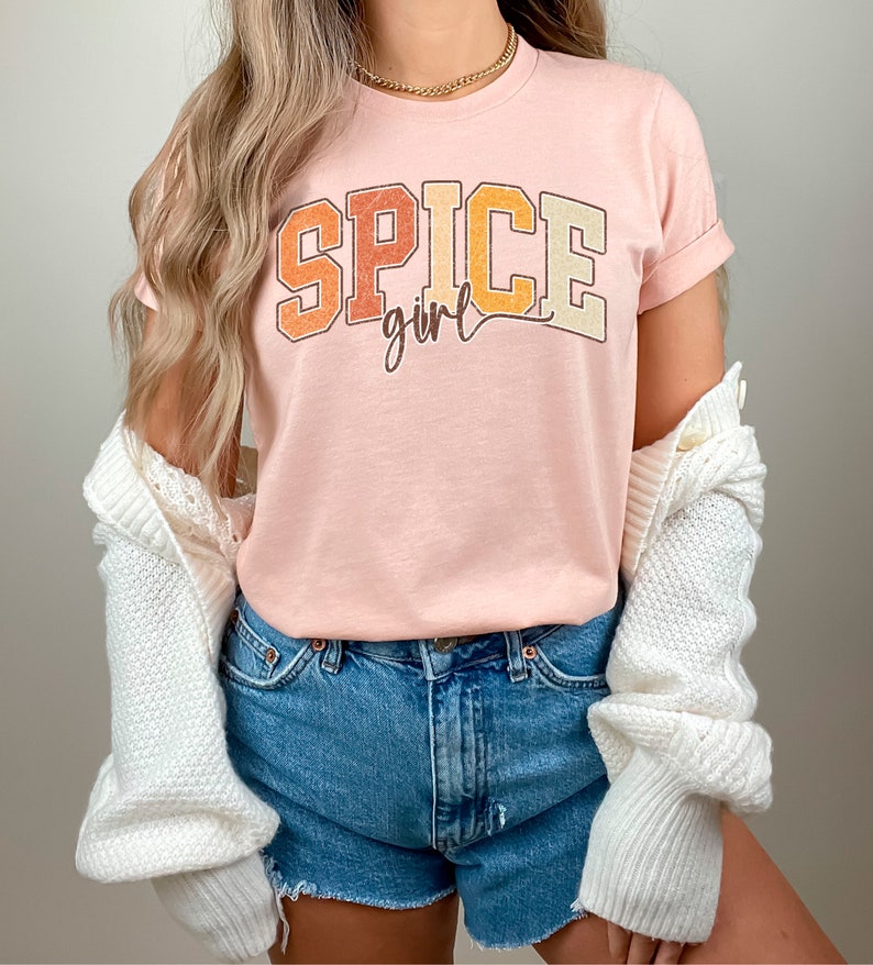 May include: A pink t-shirt with the text "SPICE girl" in a retro, colorful font. The shirt is worn with denim shorts and a white cardigan.