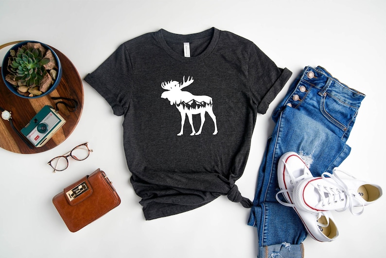 May include: Dark gray t-shirt featuring a white moose silhouette with a mountain and forest design. The shirt is styled with blue jeans and white sneakers. Other accessories include a brown wallet, glasses, and a small camera.