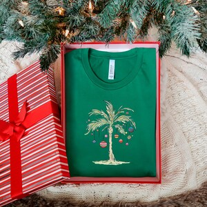 K&ouml;nnte beinhalten: Ein gr&uuml;nes T-Shirt mit einem wei&szlig;en Palmenbaum und bunten Ornamenten. Das T-Shirt befindet sich in einer rot-wei&szlig; gestreiften Geschenkbox. Der Text "Canvas" ist auf dem T-Shirt sichtbar.