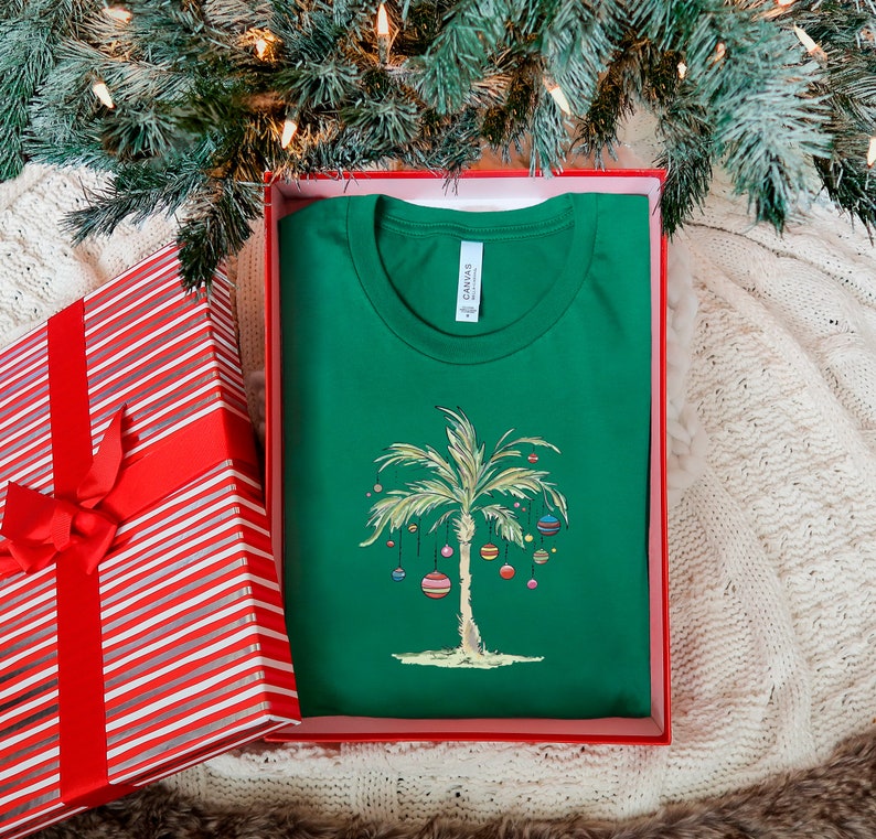 K&ouml;nnte beinhalten: Ein gr&uuml;nes T-Shirt mit einem wei&szlig;en Palmenbaum und bunten Ornamenten. Das T-Shirt befindet sich in einer rot-wei&szlig; gestreiften Geschenkbox. Der Text "Canvas" ist auf dem T-Shirt sichtbar.