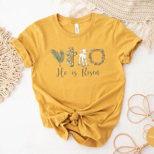 May include: Mustard yellow t-shirt with a graphic of a cross, palm branches, a lamb, and a crown of thorns. The text "He is Risen" is printed below the graphic.