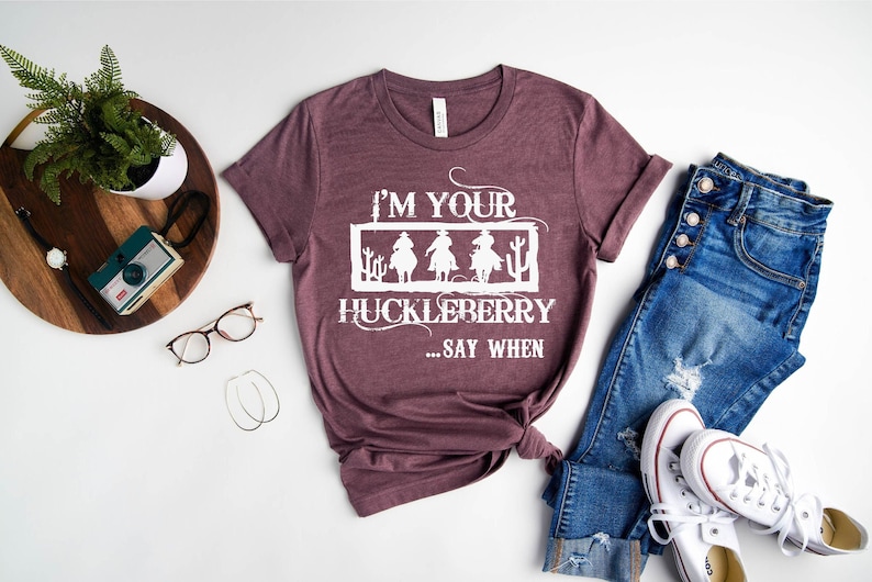 K&ouml;nnte beinhalten: Ein maronfarbenes T-Shirt mit einem wei&szlig;en Aufdruck, der "I'm your huckleberry... say when" lautet. Der Aufdruck zeigt drei Cowboys, die auf Pferden reiten. Das Shirt liegt auf einer wei&szlig;en Oberfl&auml;che mit einer Jeans und wei&szlig;en Turnschuhen.