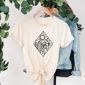 May include: A beige t-shirt with a black graphic of a desert scene with a cactus, mountains, and a sun. The text "NATURAL" is visible in the bottom right corner of the image.