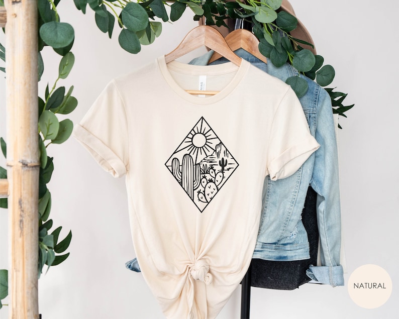 May include: A beige t-shirt with a black graphic of a desert scene with a cactus, mountains, and a sun. The text "NATURAL" is visible in the bottom right corner of the image.