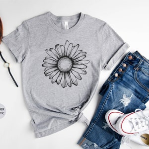 Puede incluir: Una camiseta gris jaspeada con un gr&aacute;fico de margarita en blanco y negro. La camiseta se muestra sobre un fondo blanco con un par de jeans, un reloj y un par de gafas.
