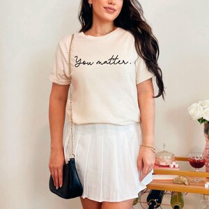 May include: A woman wearing a white t-shirt with the text "You matter." in black script. She is also wearing a white pleated skirt and a black crossbody bag.