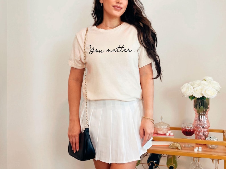 May include: A woman wearing a white t-shirt with the text "You matter." in black script. She is also wearing a white pleated skirt and a black crossbody bag.