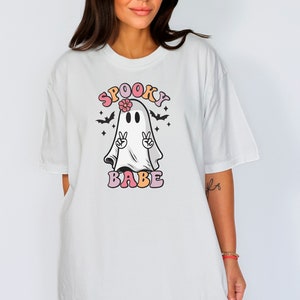 May include: White oversized t-shirt with a graphic of a ghost wearing a flower and making the peace sign. The text "SPOOKY BABE" is written in colorful letters.