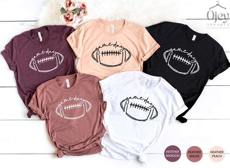 Peut inclure: Cinq t-shirts avec le texte "game day" et un graphique de football stylis&eacute;. Les t-shirts sont en marron, p&ecirc;che, blanc, noir et mauve.