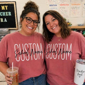以下が含まれることがあります： 「CUSTOM School Name」の白い文字が入った、お揃いの赤オレンジ色のTシャツを着た2人の女性。一人は透明なアイスドリンクを、もう一人は「teacher fuel」と書かれた白いタンブラーを持っています。「IN MY TEACHER ERA」と書かれた黒板の看板。