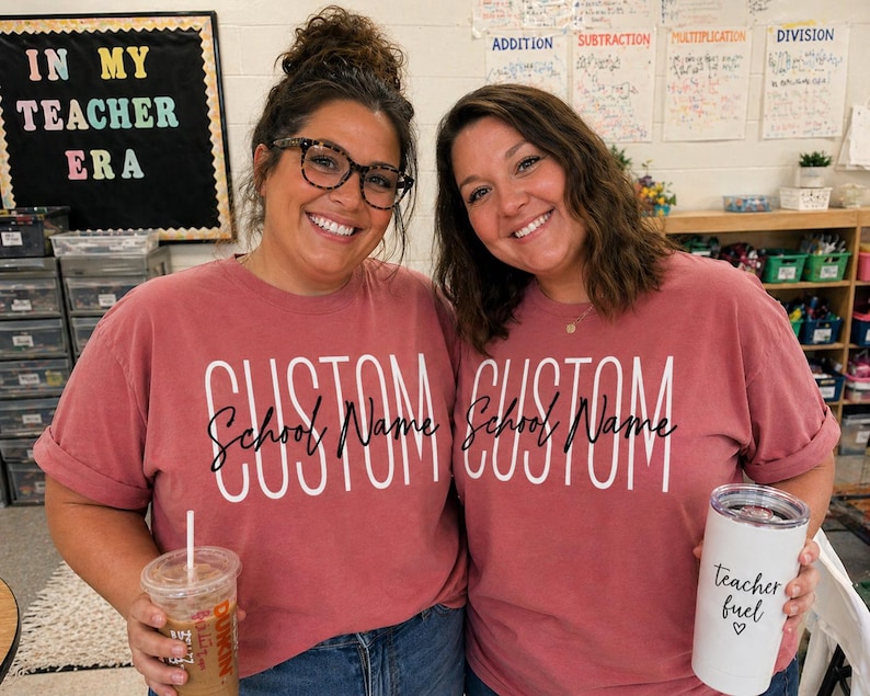以下が含まれることがあります： 「CUSTOM School Name」の白い文字が入った、お揃いの赤オレンジ色のTシャツを着た2人の女性。一人は透明なアイスドリンクを、もう一人は「teacher fuel」と書かれた白いタンブラーを持っています。「IN MY TEACHER ERA」と書かれた黒板の看板。