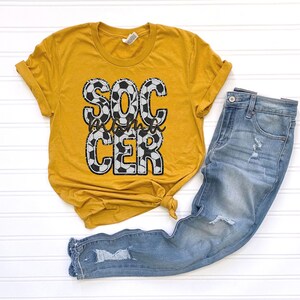 May include: A mustard yellow t-shirt with a graphic of a soccer ball pattern that reads "Soccer Mom".