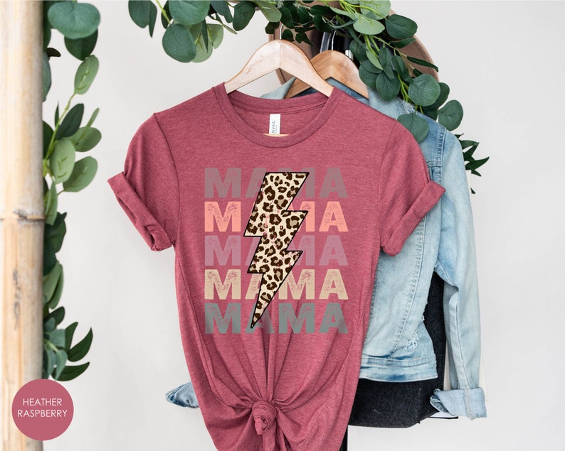 Op de afbeelding: Een heide-frambozen t-shirt met het woord "MAMA" vijf keer herhaald in een verloop van roze, oranje en grijs. Een luipaardprint bliksemgrafiek is gecentreerd op het shirt.