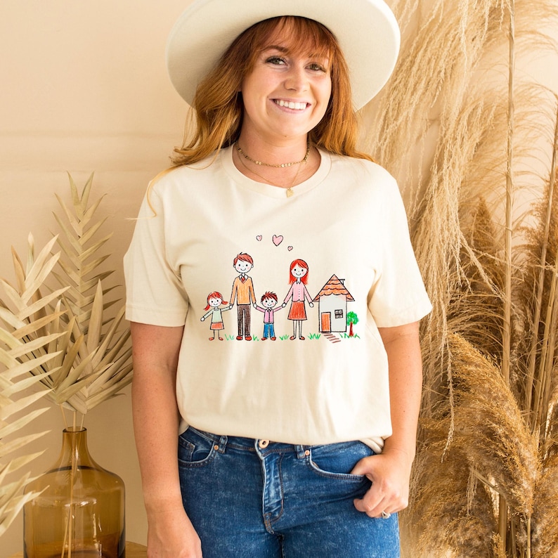 Peut inclure: Un t-shirt cr&egrave;me avec une illustration dessin&eacute;e &agrave; la main d'une famille de quatre personnes debout devant une maison. L'illustration est en rouge, orange, jaune, vert et bleu.
