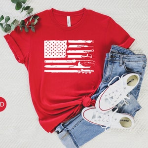 K&ouml;nnte beinhalten: Rotes T-Shirt mit einem wei&szlig;en, abgenutzten Design einer amerikanischen Flagge mit Angelruten und einem Fisch. Die Flagge ist mittig auf dem T-Shirt platziert.