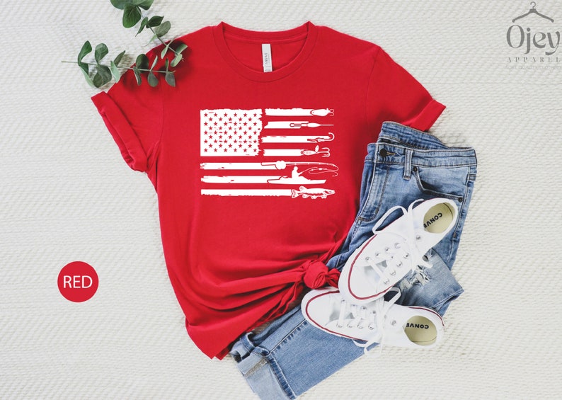 K&ouml;nnte beinhalten: Rotes T-Shirt mit einem wei&szlig;en, abgenutzten Design einer amerikanischen Flagge mit Angelruten und einem Fisch. Die Flagge ist mittig auf dem T-Shirt platziert.