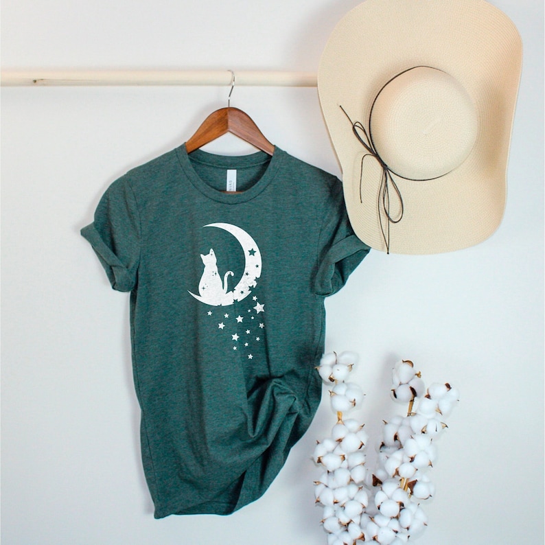 May include: A dark green t-shirt with a white graphic of a cat sitting on a crescent moon with stars.