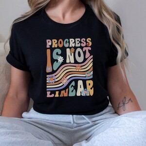 May include: Black t-shirt with the text "Progress is not linear" in a retro font with colorful stripes and flowers.