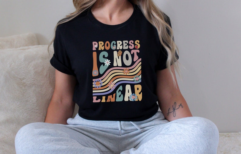 May include: Black t-shirt with the text "Progress is not linear" in a retro font with colorful stripes and flowers.