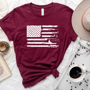 K&ouml;nnte beinhalten: Ein maroonfarbenes T-Shirt mit einem wei&szlig;en, abgenutzten Design einer amerikanischen Flagge mit Angelruten, K&ouml;dern und einem Fisch. Die Flagge ist mittig auf dem Shirt platziert.